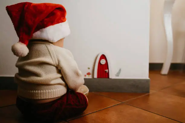 Little kid sitting near a gnomes door. He's wearing a santa hat.