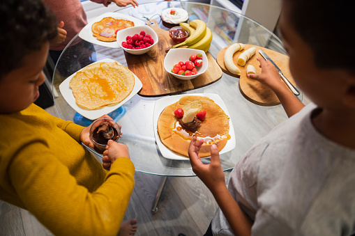 An over the shoulder shot of a young boy in the kitchen, they have baked pancakes together, they're now adding fruit and chocolate spread as tasty toppings.