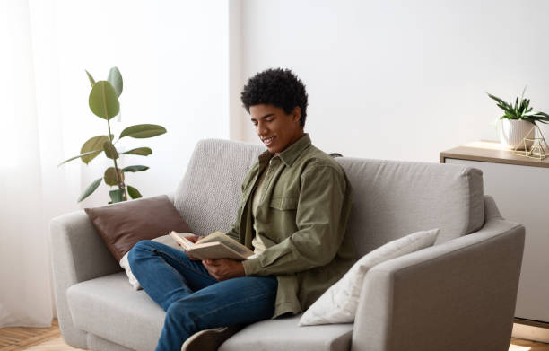 istock Cheerful black male student sitting on couch with book, reading and enjoying home studies 1290487934