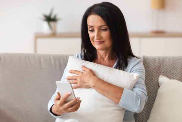 istock Smiling mature woman using mobile phone, holding pillow 1290488015