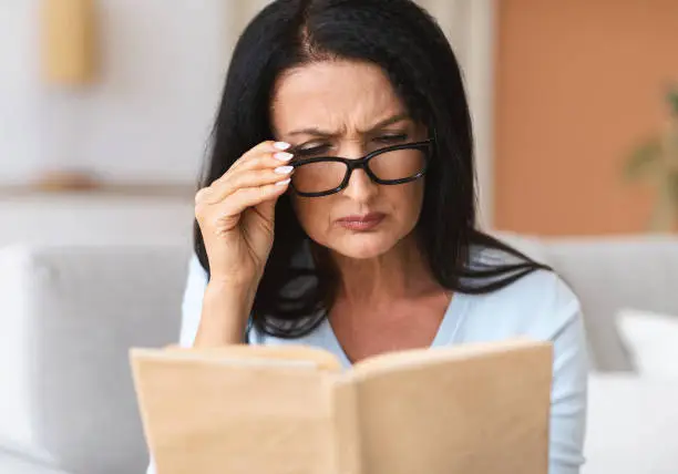 Closeup portrait of confused mature woman squinting to see more clearly, wearing and touching glasses, trying to read book, having difficulties seeing text because of vision problems, cheking diary