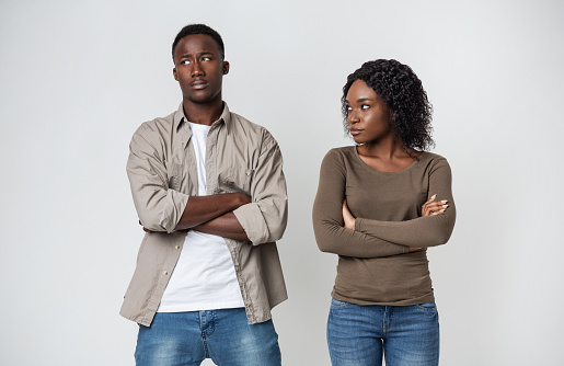 Offended black woman looking at her annoyed boyfriend, having difficulties or distrust in relationships, grey studio background. African american husband feeling cold in marriage, looking aside