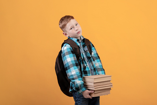 Exhausted schoolboy with backpack holding heap of books on yellow studio background, coming back home from school, copy space. Upset redhead boy have to do homework, bringing books