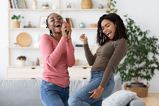 Carefree african american girlfriends singing and dancing, having fun together at home, copy space. Joyful black ladies having party, using mobile phone as microphone, living room interior