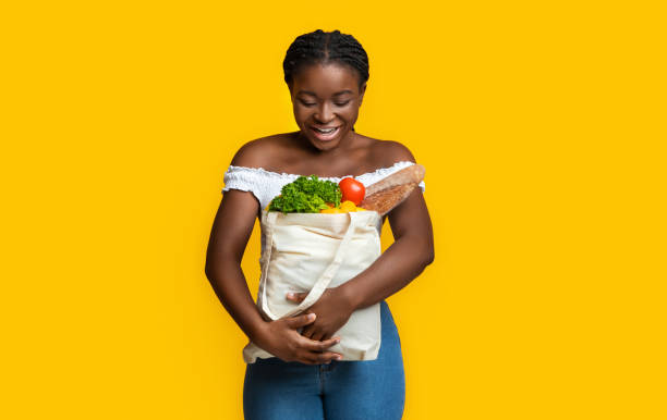 happy young black woman holding eco bag with groceries on yellow background - leaf vegetable freshness vegetable market imagens e fotografias de stock