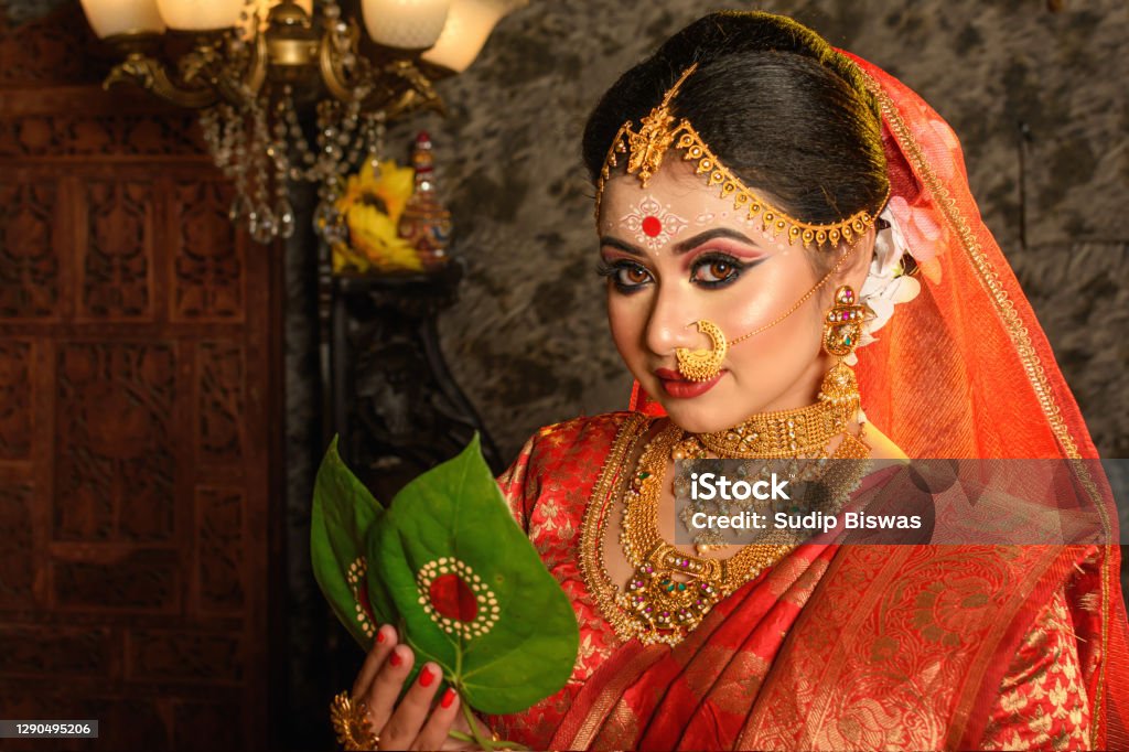 Portrait of very beautiful Indian bride holding betel leaf, Bengali bride in traditional wedding saree with makeup and heavy jewellery in studio lighting indoor Bride Stock Photo