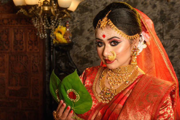 Portrait of very beautiful Indian bride holding betel leaf, Bengali bride in traditional wedding saree with makeup and heavy jewellery in studio lighting indoor stock photo