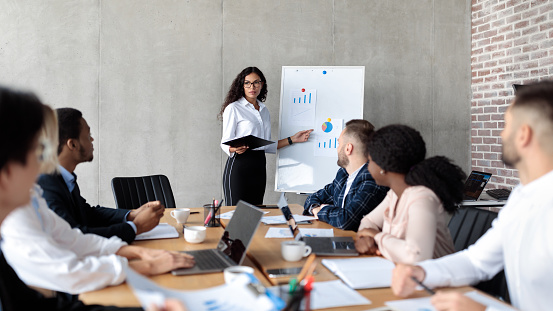 Latin Businesswoman Making Business Presentation Giving Speech For Colleagues Standing And Pointing At Blackboard With Charts And Graphs In Modern Office. Panorama, Selective Focus