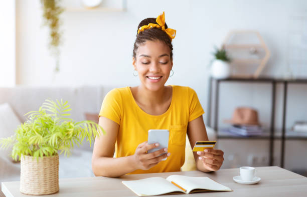 afro señora sosteniendo teléfono inteligente y tarjeta de crédito en casa - greeting card holding women credit card fotografías e imágenes de stock