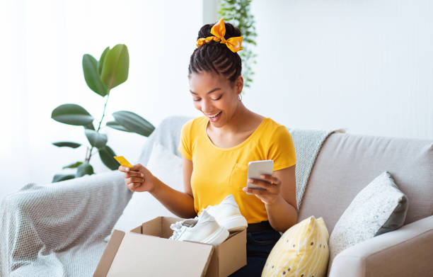 Senhora negra feliz sentada com caixa, cartão de crédito e smartphone