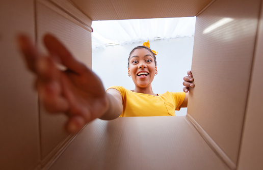 Home Delivery Service And Unboxing Concept. Happy and excited african american woman unpacking carton box and looking inside, trying to get goods outside. View from the package on overjoyed black lady