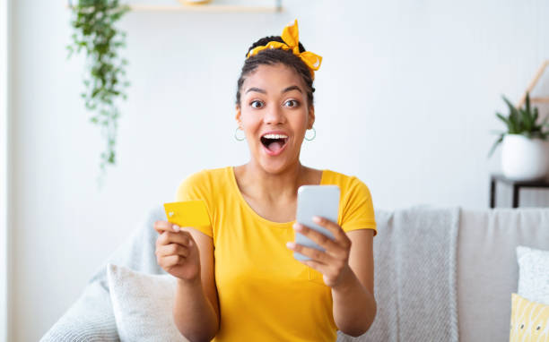 dame afro excité retenant le smartphone et la carte de crédit à la maison - internet e commerce credit card retail photos et images de collection