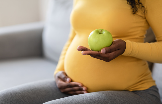 Cropped of black pregnant woman holding green apple and touching her big tummy, sitting on couch at home. Unrecognizable african american expecting lady eating fruits, copy space, closeup.