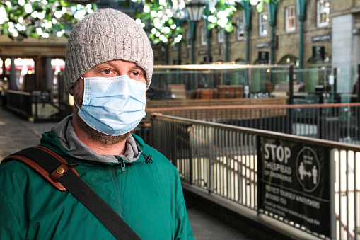 Color image depicting a caucasian male in his 30s wearing a face mask in central London during the covid-19 pandemic. In the background the Christmas decorations of Covent Garden are defocused. Room for copy space.
