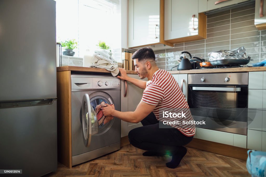 It's Laundry Day! Man crouching down to put washing into his washing machine in his kitchen.  He is in the Northeast of England. Washing Machine Stock Photo