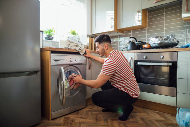 It's Laundry Day! Man crouching down to put washing into his washing machine in his kitchen.  He is in the Northeast of England. washer stock pictures, royalty-free photos & images