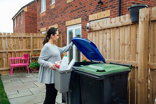 Pregnant woman disposing of her household recycling into an outdoor bin in her garden. She is in the North East of England.