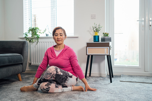 Senior woman sitting in the living room of her home doing a spinal twist yoga pose with her eyes closed. She is in the North East of England.