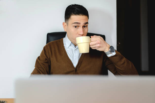 istock Man drinking a cup of hot beverage while working at computer desk with laptop. 1290520753