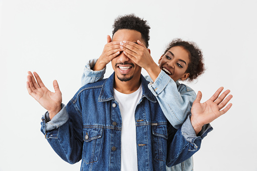 Playful african woman standing behind her boyfriend and cover his eyes over grey background