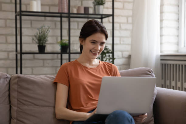 feliz mulher millennial usando computador, sentado no sofá. - relaxation working humor sofa - fotografias e filmes do acervo