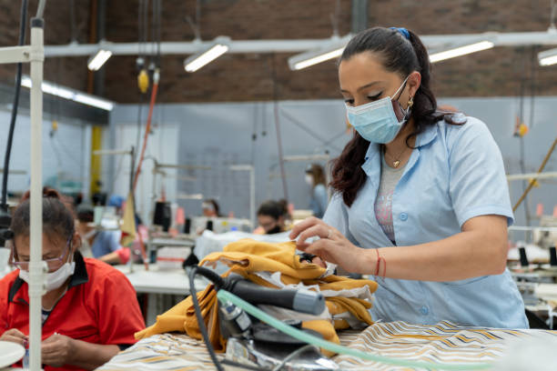 donna che lavora in una fabbrica tessile durante la pandemia indossando una maschera facciale mentre stira i vestiti - manual worker sewing women tailor foto e immagini stock