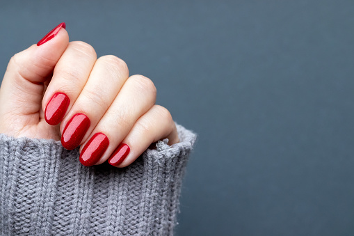 Female hand in knitted sweater with beautiful manicure - red glittered nails on gray background with copy space. Selective focus. Closeup view