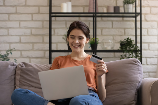 Smiling millennial european female bank client holding plastic credit card in hands, entering information cvv code in computer application, involved in online shopping, purchasing goods or services.