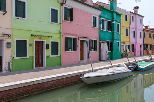 Burano, Venice, Italy - July 2, 2018: Panoramic view of brightly coloured homes and water canal with boats in Burano, it is an island in the Venetian Lagoon. People walk and rest on streets