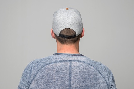 Rear view of young man wearing baseball cap standing against grey background.