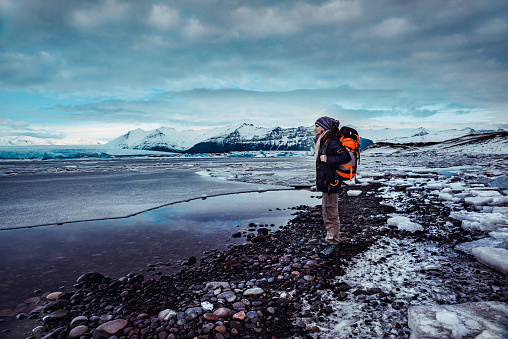 Traveling to Iceland, woman with backpack standing on the black beach and enjoying view on Jokulsarlon Glacier Lagoon, extreme winter vacation
