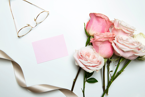 Pink paper, reading glasses, golden ribbon and roses on white table.