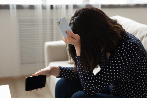 Close up unhappy frustrated Asian woman holding white empty mock up plastic card and phone, touching head, crying, having problem with credit card, loss money, internet fraud and scam concept