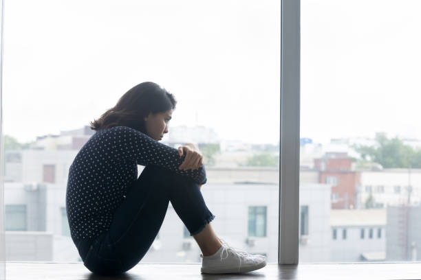 istock Side view frustrated lonely Asian woman sitting on windowsill 1290561573