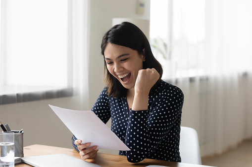 Overjoyed laughing Asian young businesswoman celebrating achievement, reading letter close up, holding paper sheet, sitting at desk, female student excited by great exam results, showing yes gesture