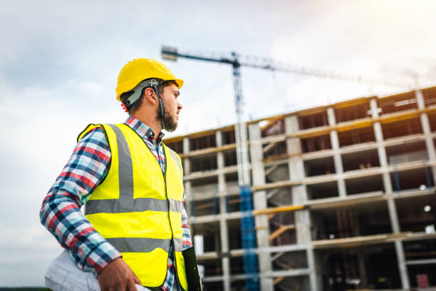 istock Young male builder/ engineer / architect / inspector / manager at construction site, holding a construction plan and looking at the construction. Architecture / construction industry concept. 1290562645