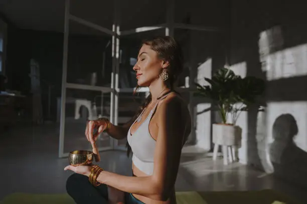 Photo of a young woman, practicing yoga and meditation, to stay healthy and calm. She is performing the exercise in her home with a Tibetan singing bowl.