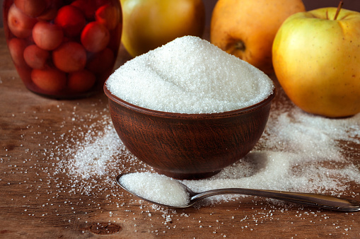 White sugar in a ceramic bowl of red clay and in a teaspoon on a wooden table with apples.