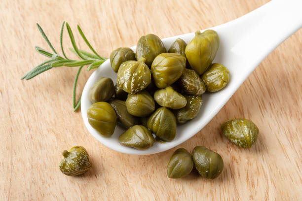 Pickled capers in a white porcelain spoon on a brown wooden cutting board. Marinated buds of caper bush. Mediterranean cuisine ingredient. Organic spices and seasonings. Pickled capers in a white porcelain spoon on a brown wooden cutting board. Marinated buds of caper bush. Mediterranean cuisine ingredient. Organic spices and seasonings. Top view. caper stock pictures, royalty-free photos & images