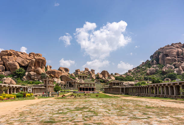 Long shot of Nandi Monolith temple in its area, Hampi, Karnataka, India. Hampi, Karnataka, India - November 4, 2013: Long wide shot of Nandi Monolith Statue temple in its area. Brown cattle grazing. Surrounded by boulders on hills under blue cloudscape. virupaksha stock pictures, royalty-free photos & images