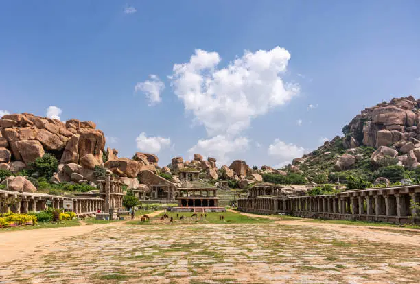 Hampi, Karnataka, India - November 4, 2013: Long wide shot of Nandi Monolith Statue temple in its area. Brown cattle grazing. Surrounded by boulders on hills under blue cloudscape.