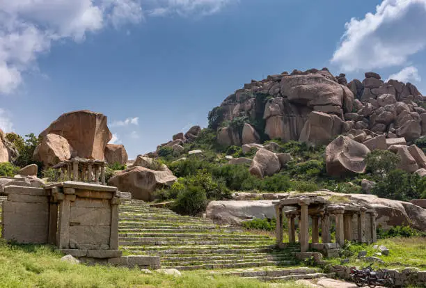 Hampi, Karnataka, India - November 4, 2013: Nandi Monolith Statue temple and area. wide stairway up on hill with ruins on side. Hill with giant boulders under blue cloudscape. Green foliage and weeds.