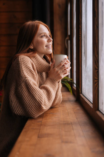 porträt von charmanten lächelnden jungen frau steht in der nähe des fensters mit tasse heißen kaffee und blick nach draußen. - winter beverage stock-fotos und bilder