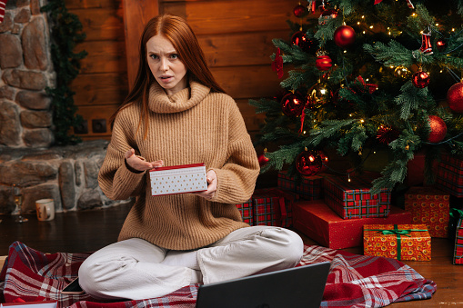 Dissatisfied young woman waiting for good Christmas present, lady with disappointment opening box with surprise on background of xmas tree at living room. Female looking at camera incomprehensibly.