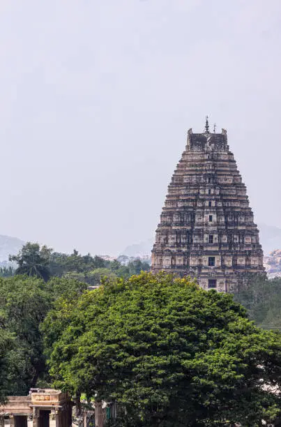 Hampi, Karnataka, India - November 4, 2013: Closeup of Virupaksha east Gopuram seen from Nandi Monolith Statue temple ruins under light blue sky. Green foliage up front. .