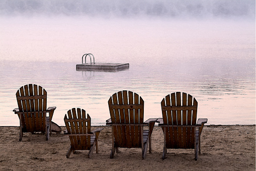 Four Adirondack chairs facing Squam Lake on a cold Fall morning with a cool magenta mist rising above the water just beyond the off shore  swimming platform. A slight water color transformation of the image gives a painterly quality to the serene scene.