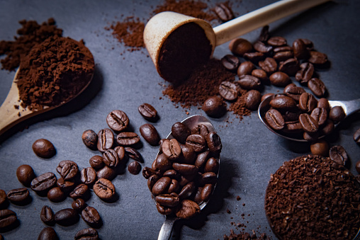 Different spoons filled with roasted coffee beans of with grinded coffee on a slate stone platter background. On the slate stone an arrangement of loose roasted coffee beans and grinded coffee is complementing the whole scenery.