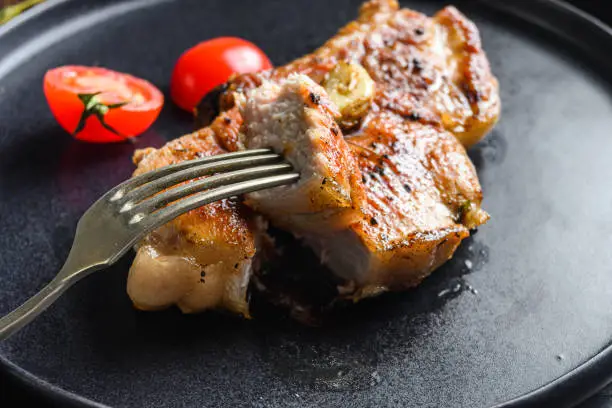 Dish of grilled pork chop with tomatoes top view with knife and slice on fork over old rustic dark wood table table close up new wide angel