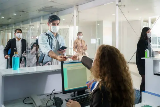 Photo of A male airline passengerwith mask is handing over his passport at the airline counter check in through an acrylic barrier for disease prevention coronavirus or covid-19 at airport for New normal travel