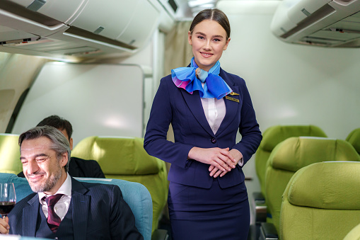 Portrait young woman in blue suit, flight attendant/air hostess in business class cabin at airplane.